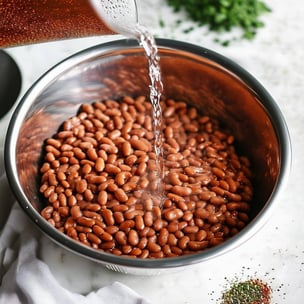 large metal bowl of dry pinto beans with water being poured in to show the process of soaking the beans as a preparation process