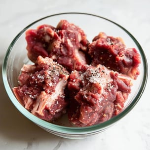raw oxtail, about 3 or 4 pieces showing the bone in a glass bowl witl salt and black pepper sprinkled on top